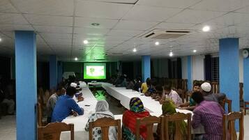 A conference room with a long table and chairs, a projector screen, and people seated facing the screen.