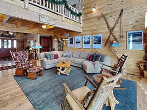 A spacious living room with a large sectional sofa, wooden chairs, and a coffee table. A balcony with a railing and a sign reading "Deep Creek Lake" is visible. - Magnificent Mountain Lodge by Taylor Made Deep Creek (McHenry)
