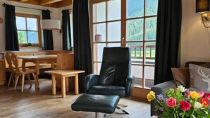A room with a wooden dining table, a black leather chair, a sofa, and a large window with curtains. - Alpine Chalet in Wald im Pinzgau for 12 (Wald im Pinzgau)