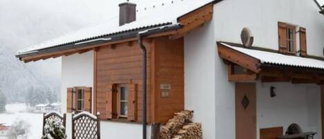 A two-story chalet with a white exterior and wooden accents, a covered porch, and a stack of firewood outside.