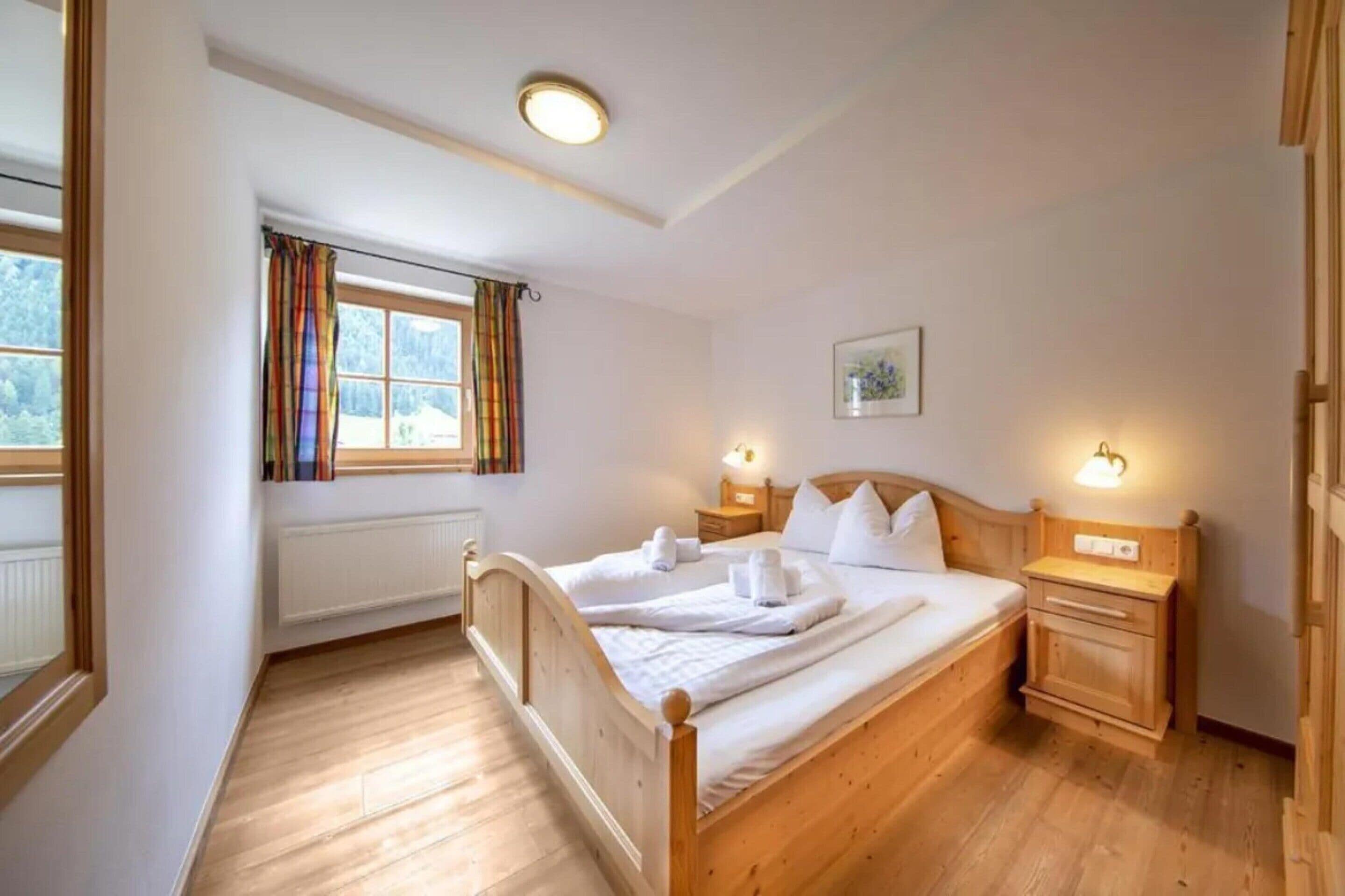 Ein Schlafzimmer mit einem hölzernen Bett, einem Fenster mit Vorhängen, einem Nachttisch mit Lampe sowie einem an der Wand hängenden Bild.