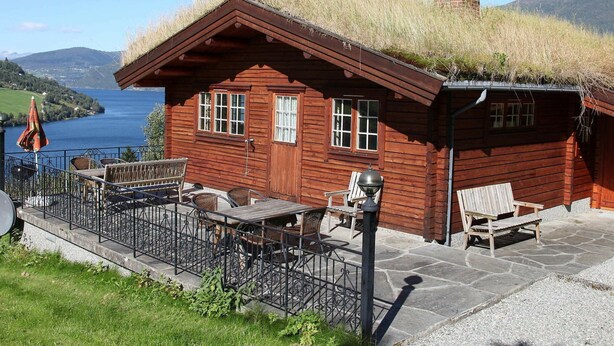 Une cabane en bois avec un toit de chaume, une terrasse en pierre et une vue sur un lac.
