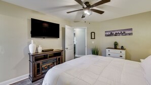 A bedroom with a large bed, a ceiling fan, a flat-screen TV mounted on the wall, a dresser, and a door leading to another room.