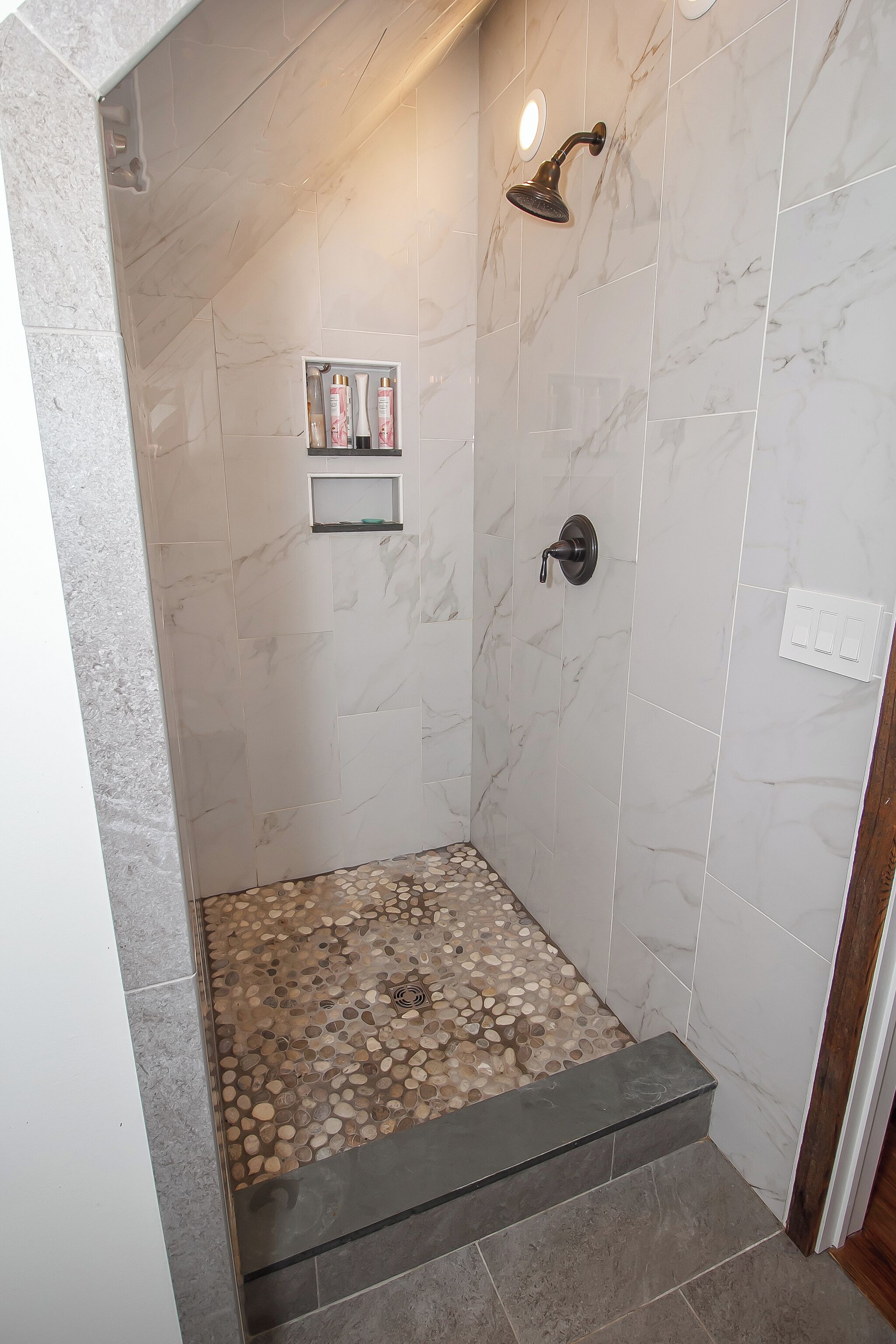 Ducha con pared de mármol, estante empotrado con artículos de aseo y suelo con diseño de guijarros.