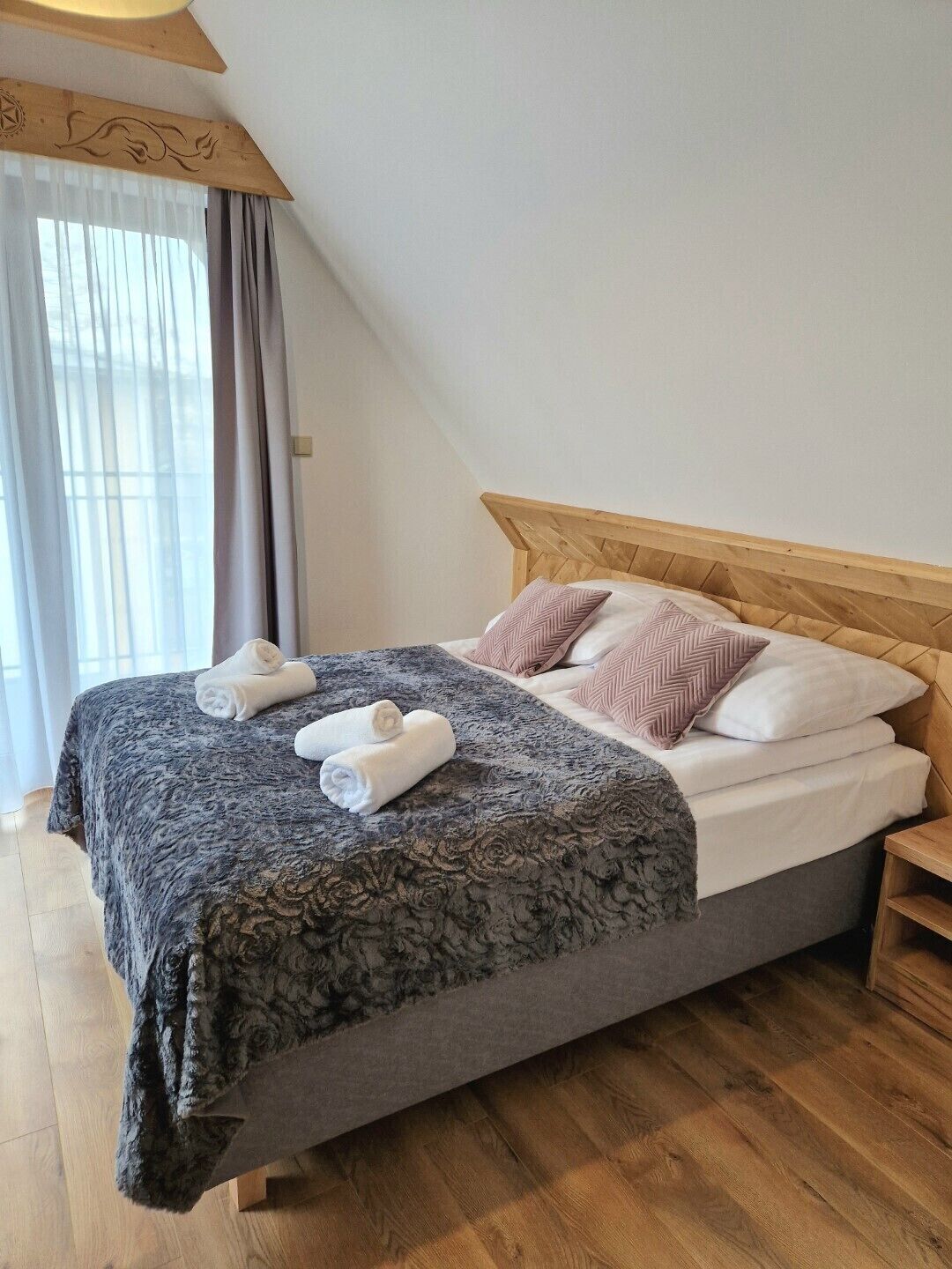 A neatly made bed with a grey patterned bedspread and two pillows, a wooden headboard, and a wooden nightstand with a drawer.
