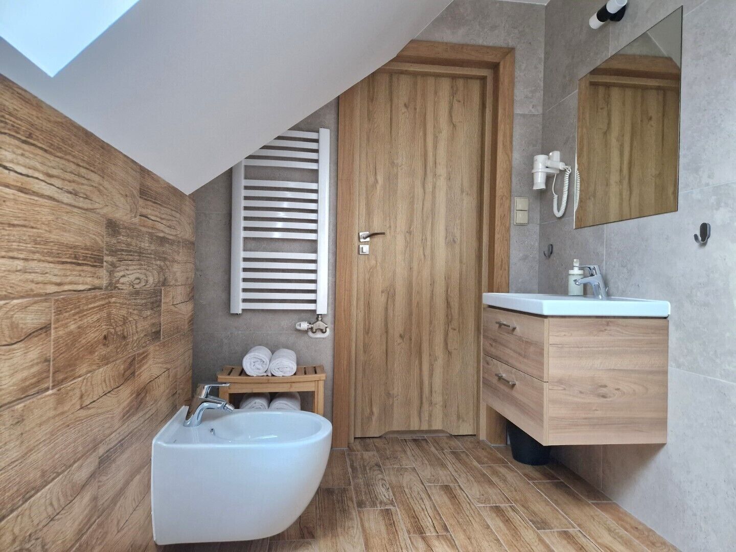 A modern bathroom with a freestanding bathtub, wooden cabinetry, a large mirror, and a skylight.