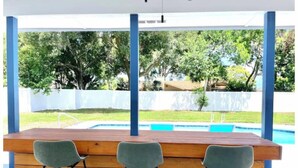 Un comptoir en bois, avec trois tabourets de bar gris, donnant sur une piscine et un espace verdoyant.