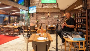 Un hombre tocando la guitarra en un restaurante con paredes de ladrillo, muebles de madera y varios televisores en la pared.