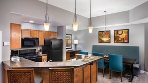 Une cuisine moderne dotée de plans de travail en granit, d’armoires en bois, d’un four à micro-ondes encastré et d’un réfrigérateur. On y trouve également un coin repas avec des chaises bleues et une petite table.