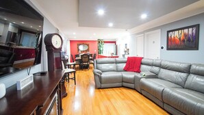Un salón espacioso con un sofá de cuero gris, suelo de madera, una zona de comedor con una pared de acento roja y un gran reloj en la pared.