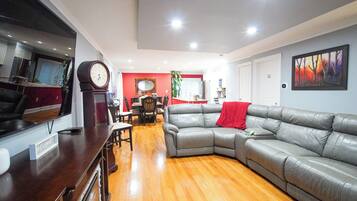 A spacious living room with a grey leather sofa, wooden flooring, a dining area with a red accent wall, and a large clock on the wall.