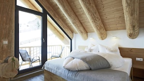 Ein Schlafzimmer mit Holzbalkendecke, einem großen Fenster, einem Bett mit weißer und grauer Bettwäsche und einem Sessel am Fenster.