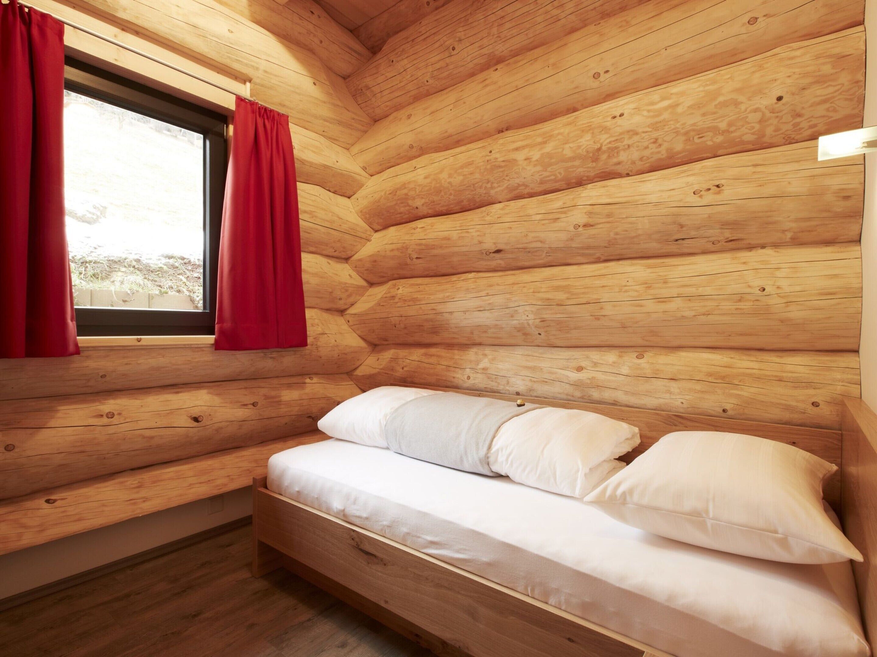 A log cabin room with a bed, red curtains, and a window.