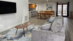 A living room with a grey sofa, a coffee table, and a TV mounted on the wall. There is a dining area with a wooden table and chairs, and a kitchen in the background. - POOL House+Garage. Bring your Bikes! + 3Bed/2Bath
 (Port Orange)