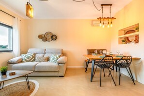 A modern living room with a sofa, dining table, and chairs. There is a window, a hanging light fixture, and wall decor. - Cozy apartment in charming Costa da Caparica with AC, WiFi (Costa da Caparica)