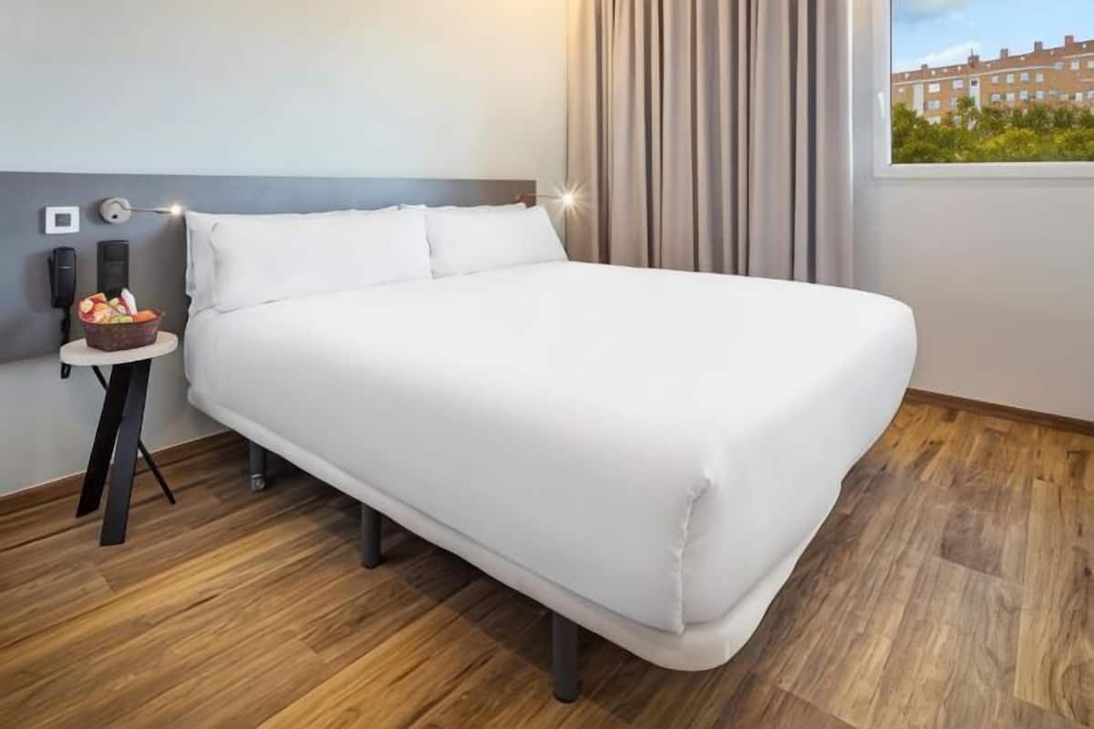 Un lit bien fait dans une chambre d’hôtel, avec un plancher en bois, une petite table sur laquelle se trouve un panier de fruits, et une fenêtre avec des rideaux.