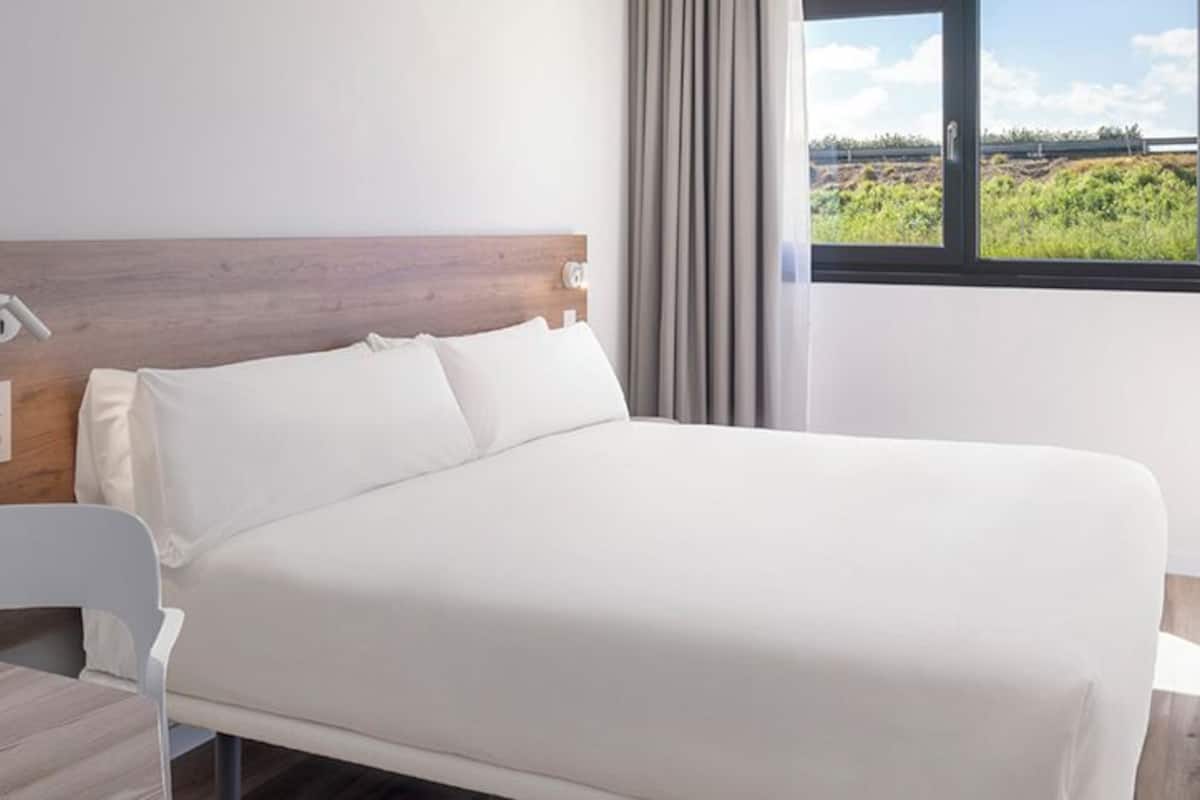 Une chambre d’hôtel dotée d’un grand lit, d’une tête de lit en bois, d’un bureau avec une chaise, d’une fenêtre donnant sur un paysage verdoyant et d’un téléviseur à écran plat fixé au mur.