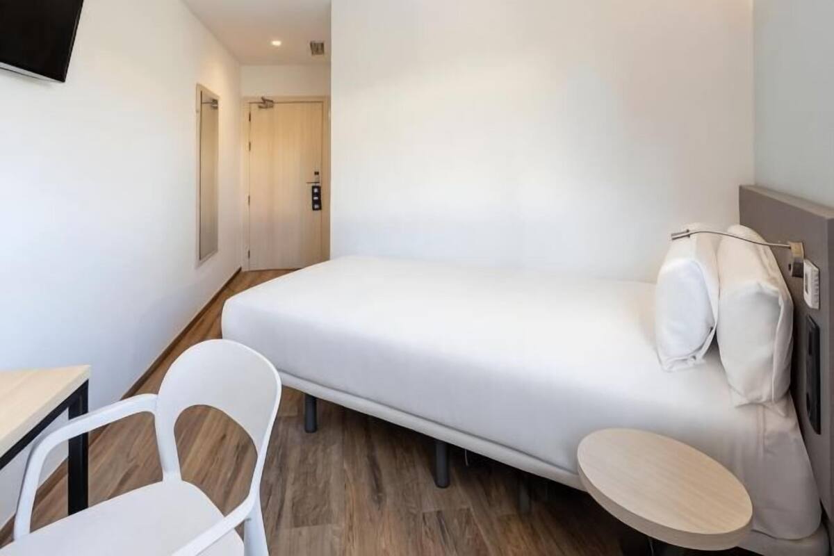Une petite chambre moderne, équipée d’un lit, d’une chaise, d’une petite table et d’une porte donnant sur une autre pièce.