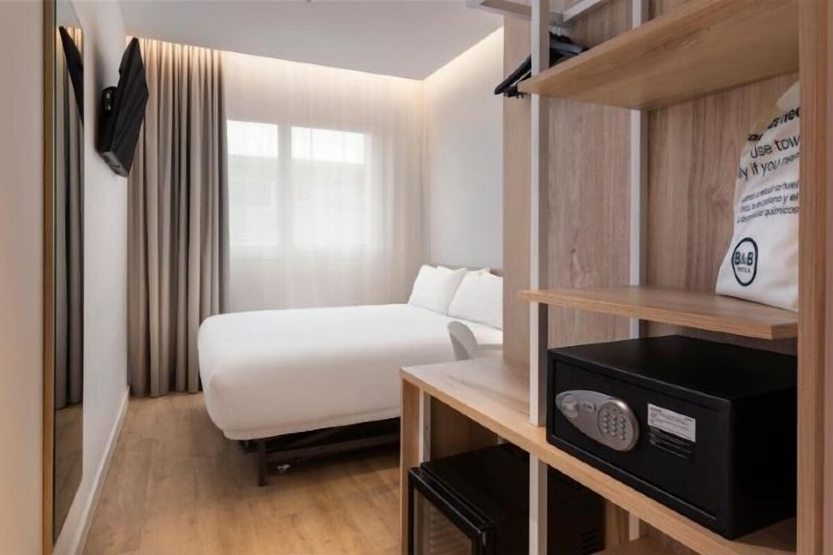 Ett hotellrum med en säng, ett skrivbord, en stol, en tv och en hylla med en väska och en skylt.