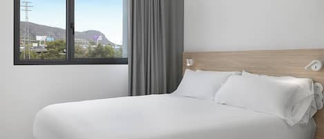 A hotel room with a large bed, a window with a view of buildings and hills, and a wooden headboard.