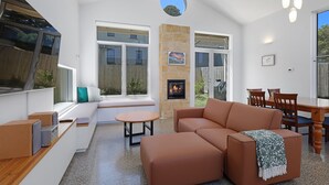 A modern living room with a fireplace, a dining area, and a large window overlooking a garden. - The Sunshine House - Nestled in the heart of the city. (Warrnambool)
