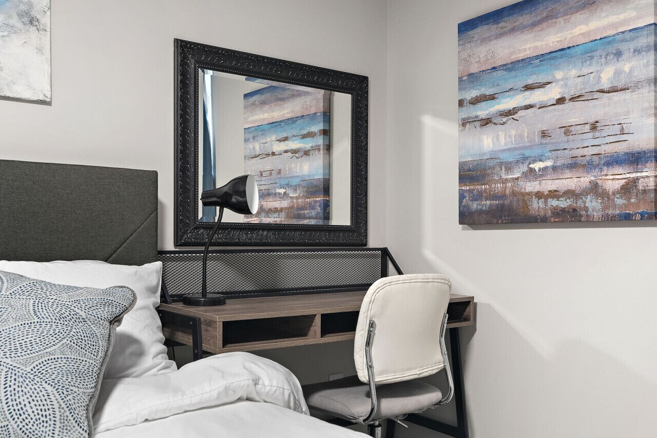 A bedroom with a bed, a desk, a chair, a mirror, and two paintings on the wall.