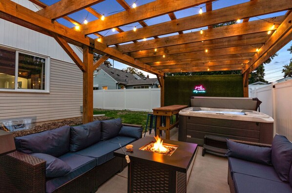 An outdoor patio with a wooden pergola, a fire pit, a hot tub, and a seating area with cushions.