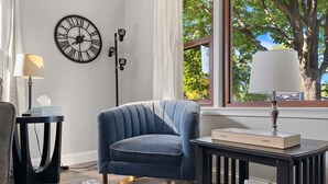 A room with a blue armchair, a round wall clock, a lamp, a small table with a book, and a window with a view of trees. - Ultimate Group Getaway: Sleeps 16, Hot Tub, Arcade, & 5 Min from Mall of America (Minneapolis)