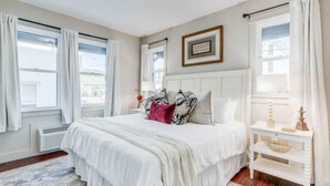 A bedroom with a large bed, white curtains, a bedside table with a lamp, and a framed picture on the wall.