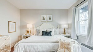 Une chambre à coucher avec un grand lit, deux tables de chevet avec des lampes, une fenêtre avec des rideaux et des tableaux encadrés accrochés au mur.