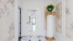 Un couloir doté d’un sol en marbre, de murs blancs ornés de motifs floraux, d’une porte blanche et d’une colonne sur laquelle est posée une plante.