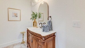 A bathroom with a wooden vanity, a large mirror, and a small round table. - The Golden Hour Room (Manteo)