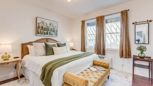 A bedroom with a bed, bedside tables, a bench, a window with curtains, a mirror, and artwork on the wall. - The Golden Hour Room (Manteo)