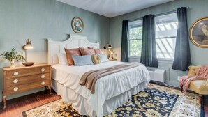 A bedroom with a bed, bedside table, lamp, and a large window with curtains. - The Mozart's Hideaway Room (Manteo)