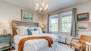 A bedroom with a large bed, a wooden nightstand, a chair, a window with curtains, and a chandelier. - The Harbor Haven Room (Manteo)