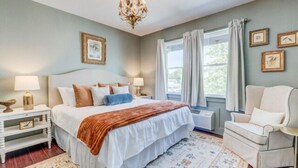 A bedroom with a large bed, a chandelier, a window with curtains, and a chair. - The Poets Sanctuary Room (Manteo)
