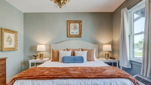 A neatly arranged bedroom with a large bed, two bedside tables with lamps, a framed picture on the wall, and a window with curtains. - The Poets Sanctuary Room (Manteo)