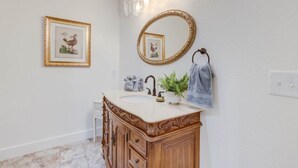 A bathroom with a wooden vanity, a round mirror, and framed pictures on the wall. - The Poets Sanctuary Room (Manteo)