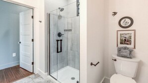 A bathroom with a glass shower enclosure, a toilet, and a door leading to another room. - The Poets Sanctuary Room (Manteo)