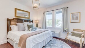 A bedroom with a large bed, a bedside table with a lamp, a window with curtains, a chair, and a framed picture on the wall.