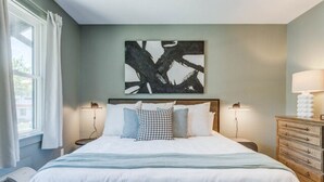 A bedroom with a large bed, two bedside tables, a dresser, and a wall-mounted abstract painting. - The Fly South - Room (Manteo)