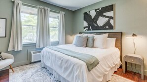 A bedroom with a large bed, a bedside table, a window with curtains, and a wall-mounted artwork. - The Fly South - Room (Manteo)