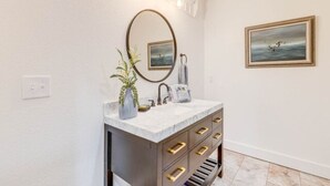 A bathroom with a vanity, a mirror, a towel rack, and a framed picture on the wall. - The Fly South - Room (Manteo)