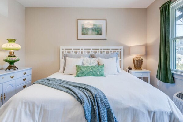 A neatly made bed with a white headboard, a green throw blanket, and multiple pillows. There is a framed picture on the wall above the bed, and a window with green curtains to the right.