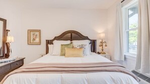 A neatly made bed with a wooden headboard, a bedside table with a lamp, and a window with curtains. - The Blushing Rose Room (Manteo)