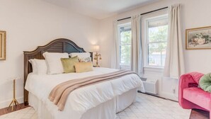 A bedroom with a large bed, a bedside table, a lamp, a window with curtains, and a pink chair. - The Blushing Rose Room (Manteo)