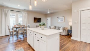 A modern kitchen with wooden flooring, a central island, pendant lighting, and a dining area with chairs and a table. - Soundscape Family Suite/pet-friendly (Manteo)