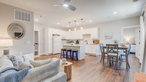 A modern kitchen with white cabinets, a central island with stools, and a dining area with wooden chairs. - Soundscape Family Suite/pet-friendly (Manteo)