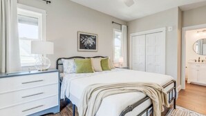 A bedroom with a bed, bedside table, and a nightstand. There is a window with curtains, a ceiling fan, and a door leading to another room. - Soundscape Family Suite/pet-friendly (Manteo)