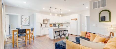 A modern living room with a sofa, a dining table with chairs, a kitchen area with white cabinets, and a ceiling fan.